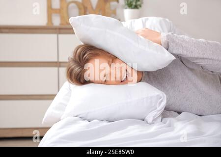 Hübscher junger, unzufriedener Mann im Pyjama, der zu Hause Ohren mit Kissen auf dem Bett zustopft Stockfoto