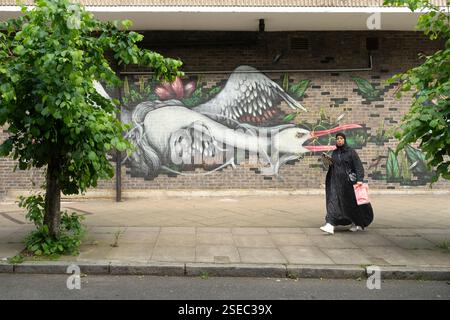 Frau läuft entlang einer Straße in der Chrisp Street, Poplar, East London, Großbritannien, in der Nähe eines Gebäudes mit einem Wandgemälde eines Storchs. Ein lebendiges Streetart-Stück. Stockfoto