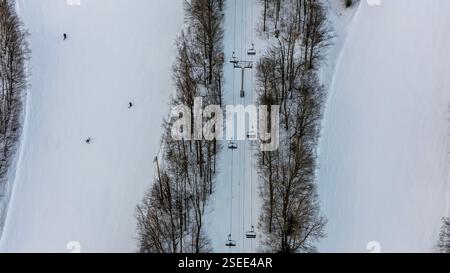 Ein Blick aus der Vogelperspektive fängt einen Skilift ein, der über einen schneebedeckten Bergort gleitet. Skifahrer fahren frische Pfade unter sich, umgeben von hohen Kiefern und maj Stockfoto