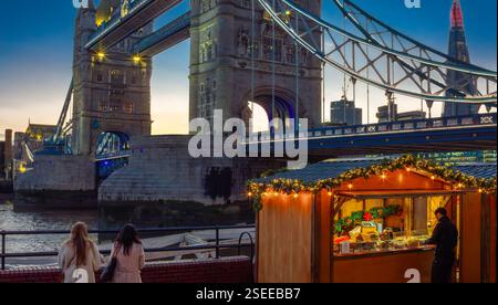 London, Großbritannien - 24. November 2024: Weihnachtsmarkt in der Nähe der Tower Bridge in der Abenddämmerung, mit zwei Personen, die die Aussicht bewundern. Der Stand ist dekoriert Stockfoto