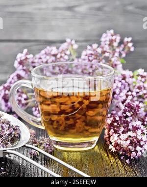 Oregano Kräutertee in einer Glasschale, frische Blumen und Metallsieb mit getrockneten Majoran-Blüten auf Holzbrett Stockfoto
