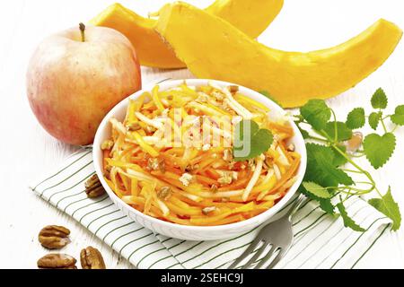 Kürbis-, Karotten- und Apfelsalat mit Pekannüssen, gewürzt mit Pflanzenöl in einer Schüssel auf einem Handtuch, Minze auf Holzbrett Hintergrund Stockfoto