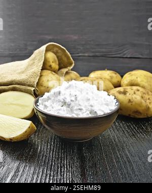 Kartoffelstärke in Tonschüssel, Gemüseknollen in einem Beutel und auf dem Tisch vor dem Hintergrund eines schwarzen Holzbretts Stockfoto