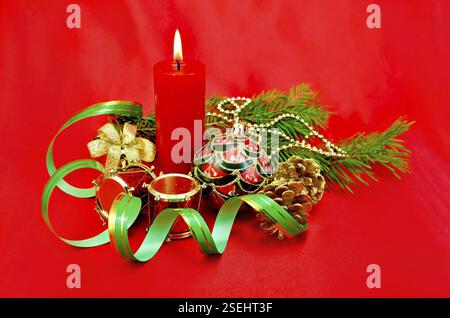 Rote brennende Kerze, Weihnachtsbaumschmuck in Form einer Kugel, eine Handglocke mit Schleife, eine Trommel, Kegel auf roter Seide Stockfoto