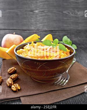 Kürbis-, Karotten- und Apfelsalat mit Pekannüssen, gewürzt mit Pflanzenöl in einer Schüssel auf einem Handtuch, Minze auf schwarzem Holzbrett Hintergrund Stockfoto