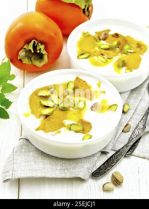 Dessert aus Joghurt, Persimon und Honig mit Vanille, Kardamom und Pistazien in zwei Schüsseln auf einem Handtuch, Minze und Löffel auf Holzbrett Hintergrund Stockfoto