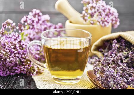 Oregano Kräutertee in einer Glastasse auf einer Quappe Serviette, frische Blumen in Mörser und auf dem Tisch, getrocknete Majoran Blumen in Beutel und Löffel auf Holzschwein Stockfoto
