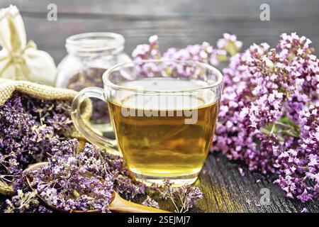 Oregano Kräutertee in einer Glasschale, frische Blumen, getrocknete Majoran Blumen in einer Tasche, Glas und Löffel auf Holzbrett Hintergrund Stockfoto