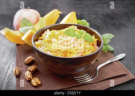 Kürbis-, Karotten- und Apfelsalat mit Pekannüssen, gewürzt mit Pflanzenöl in einer Schüssel auf einer Serviette, Minze auf dunklem Holzbrett Hintergrund Stockfoto