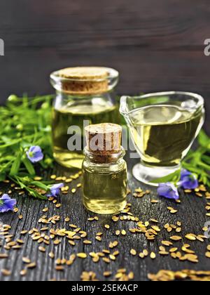 Leinöl in zwei Gläsern und ein Saucenboot mit Samen, Blättern und Leinenblumen auf schwarzem Holzbrett Hintergrund Stockfoto