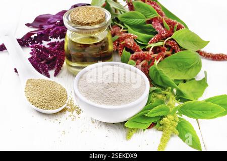 Amaranth-Mehl in einer Schüssel, Samen in einem Löffel und Öl in einem Glas, braune, grüne und violette Blüten einer Pflanze auf dem Hintergrund von hellem Holzbrett Stockfoto