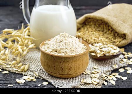 Mehl von Hafer in eine Schüssel geben, die Milch in einer Kanne, Haferflocken in einem Löffel auf Sackleinen, Korn in der Tasche, Vollkorn-hafer Stiele auf Holzbrett Hintergrund Stockfoto