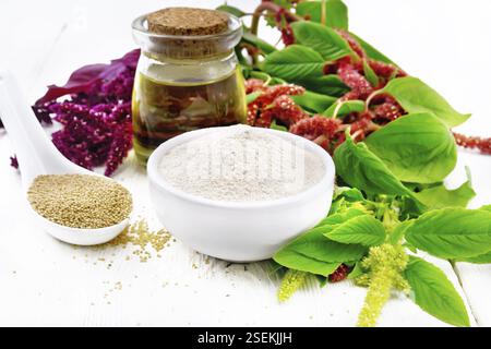Amaranth-Mehl in einer Schüssel, die Samen im Löffel und das Öl im Glas, braune, grüne und violette Blüten einer Pflanze auf dem Hintergrund des Holzbretts Stockfoto