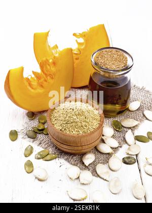 Kürbismehl in der Schüssel und Öl im Glas auf der Burlap, die Samen auf dem Tisch und die Scheiben des Gemüses auf dem Hintergrund des hellen Holzbretts Stockfoto