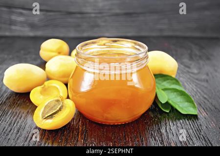 Marmelade Aprikose in Glas, Früchte und ein Zweig mit grünen Blättern auf dem Hintergrund der dunklen Holzplatte Stockfoto