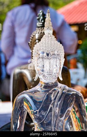 Transparente buddha-Statue aus Glas in Meditationshaltung zwischen verschiedenen buddha-Statuen auf dem unscharfen Hintergrund von Menschen und Gebäuden in Thailands Temp Stockfoto