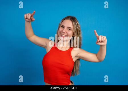 Fröhliche junge Frau, die mit beiden Zeigefingern seitwärts zeigt, lächelt und ein rotes Oberteil auf blauem Hintergrund trägt Stockfoto