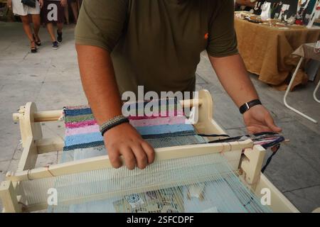 Ein Mann webt mit einem kleinen Webstuhl auf einem Flohmarkt. Er konzentriert sich auf die Aufgabe. Der Webstuhl hält Fäden in Blau- und Rosa-Tönen. Stockfoto