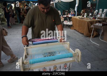 Ein Mann webt mit einem kleinen Webstuhl auf einem Flohmarkt. Er konzentriert sich auf die Aufgabe. Der Webstuhl hält Fäden in Blau- und Rosa-Tönen. Stockfoto