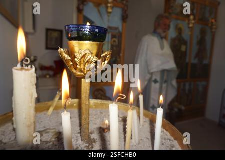 Zündete Kerzen stehen auf einer runden Plattform in einer Kirche. Ein dekorativer goldener Kerzenhalter mit blauem Halter befindet sich inmitten der Kerzen neben einem Priester Stockfoto