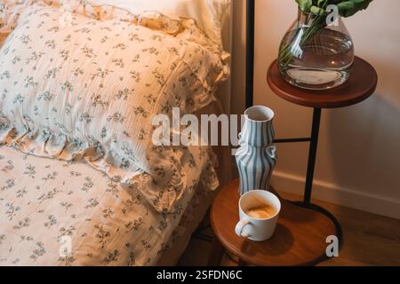 Innenraum des Schlafzimmers mit einer Tasse Kaffee auf einem Nachttisch, einer Vase mit Blumen und einer beleuchteten Lampe neben einem Doppelbett im Schlafzimmer Stockfoto