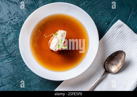 Suppe oder Consomme, dicke Fleischbrühe mit Karotten, Hühnerknödel und Mikrogrün, serviert auf einem weißen Teller auf dunkelgrünem Hintergrund mit Serviette A Stockfoto