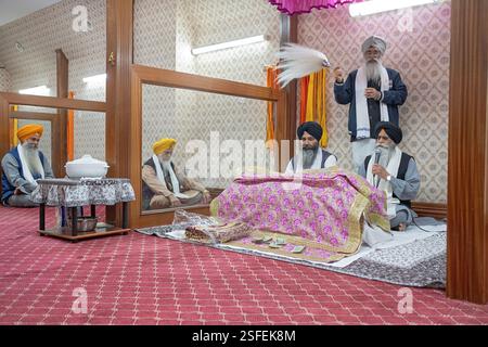 Bei einem Morgengottesdienst in einem Sikh-Tempel lasen Priester ihr heiliges Buch Guru Granth Shaib, während ein dritter Mann respektvoll einen weißen Chaur Sahib winkt. In Queens Stockfoto