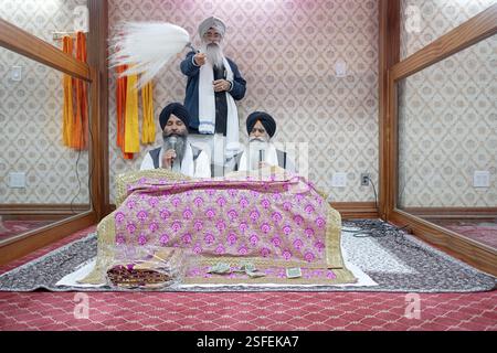 Bei einem Morgengottesdienst in einem Sikh-Tempel lasen Priester ihr heiliges Buch Guru Granth Shaib, während ein dritter Mann respektvoll einen weißen Chaur Sahib winkt. In Queens Stockfoto