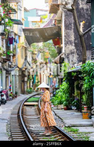 Eine Frau in einem Blumenkleid und einem traditionellen vietnamesischen konischen Hut steht auf den Spuren von Hanois berühmter Train Street Stockfoto