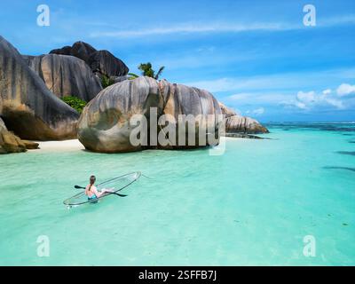 Eine Frau paddelt mit einem transparenten Kajak in der Nähe der riesigen Granitfelsen von Anse Source d'Argent auf La Digue, Seychellen, mit ruhigem türkisfarbenem Wasser Stockfoto
