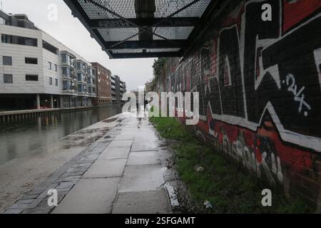 Menschen laufen entlang des Trawpath des Regents Canal, London, vorbei an einer roten Backsteinmauer, die mit Graffiti bedeckt ist Stockfoto