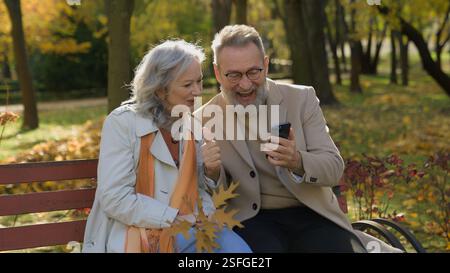 Fröhliches Paar 60er Jahre alte Frau Mann mit Handy auf Bank im Herbstpark mittleren Alters Ehefrau glücklich feiern gute Nachrichten Sieg reif Stockfoto