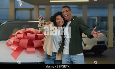 Glückliches lächelndes Paar Afroamerikaner Familienfrau Mann neue Autobesitzer zeigen Schlüssel vor der Kamera und kaufen Automobil mit großem Bogen im Händlerladen Stockfoto