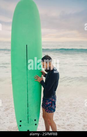 Junger bärtiger Mann mit Surfbrett, der in der Nähe eines Strandes steht. Mann mit Surfbrett im Freien an einem Sommertag. Stockfoto