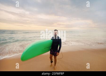 Junger bärtiger Mann mit Surfbrett, der in der Nähe eines Strandes steht. Mann mit Surfbrett im Freien an einem Sommertag. Stockfoto
