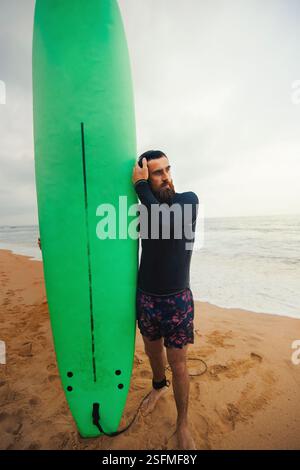Junger bärtiger Mann mit Surfbrett, der in der Nähe eines Strandes steht. Mann mit Surfbrett im Freien an einem Sommertag. Stockfoto