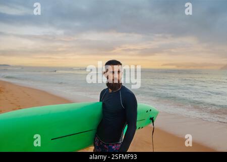 Junger bärtiger Mann mit Surfbrett, der in der Nähe eines Strandes steht. Mann mit Surfbrett im Freien an einem Sommertag Stockfoto