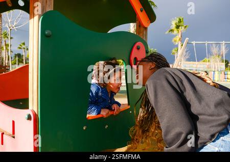 Mutter und Tochter genießen die Zeit zusammen und spielen Verstecken auf der farbenfrohen Spielplatzrutsche Stockfoto