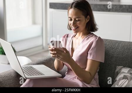 Die junge Frau sitzt bequem auf dem Sofa mit Laptop und Smartphone Stockfoto