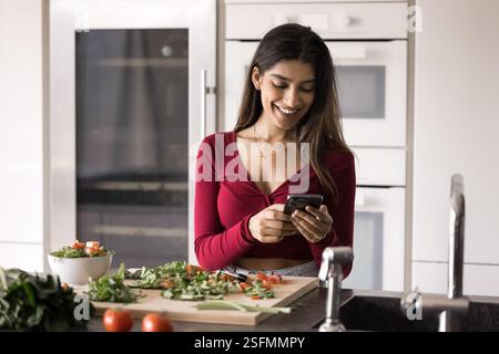 Junge Indianerin steht in moderner Küche mit Smartphone Stockfoto
