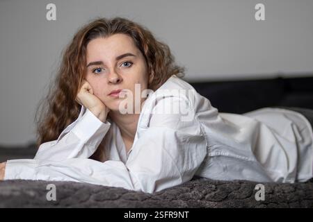 Die junge Frau liegt auf einem Bett, trägt ein weißes Hemd, den Kopf auf der Hand, mit einem kontemplativen Ausdruck Stockfoto