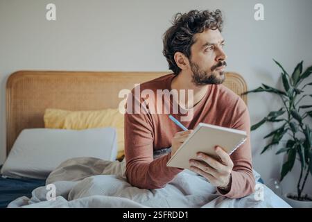 Junger, gutaussehender Mann aus dem Nahen Osten, der im Bett sitzt und in seinem Notizbuch schreibt. Kreativität und Reflexion am Morgen. Stockfoto