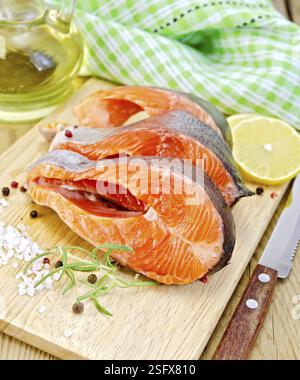 Drei Stücke Forelle mit Rosmarin und grobem Salz, Pfeffer, Messer, Pflanzenöl, Zitrone, grünes Tuch auf einem Holzbrett Hintergrund Stockfoto
