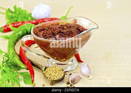 Tabasco Adjika in einem Glas Soße Boot, heißer Pfeffer, Knoblauch, Petersilie, Bockshornklee auf dem Hintergrund eines groben Stoffsacks Stockfoto