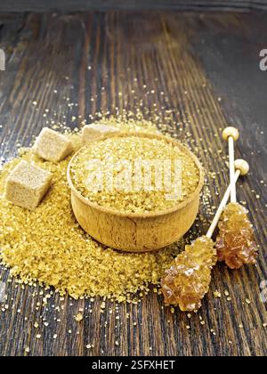 Der körnige braune Zucker in der Schüssel und auf dem Tisch, kristallin auf dem Stock und die Zuckerwürfel auf dem hölzernen Brett Hintergrund Stockfoto