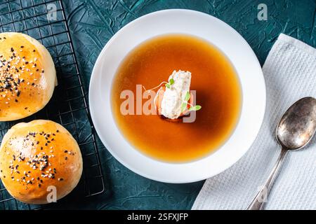 Suppe oder Consomme, dicke Fleischbrühe mit Karotten, Hähnchenknödel serviert auf einem weißen Teller auf dunkelgrünem Hintergrund mit Sesambrötchen, Serviette und Stockfoto