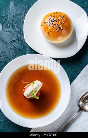 Suppe oder Consomme, dicke Fleischbrühe mit Karotten, Hähnchenknödel serviert auf einem weißen Teller auf dunkelgrünem Hintergrund mit Sesambrötchen, Serviette und Stockfoto