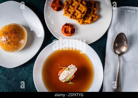 Suppe oder Consomme, dicke Fleischbrühe mit Karotten, Hähnchenknödel serviert auf einem weißen Teller auf dunkelgrünem Hintergrund mit Sesambrötchen, Serviette und Stockfoto