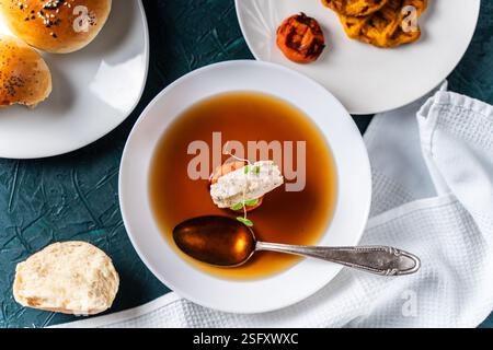 Suppe oder Consomme, dicke Fleischbrühe mit Karotten, Hähnchenknödel serviert auf einem weißen Teller auf dunkelgrünem Hintergrund mit Sesambrötchen, Serviette und Stockfoto