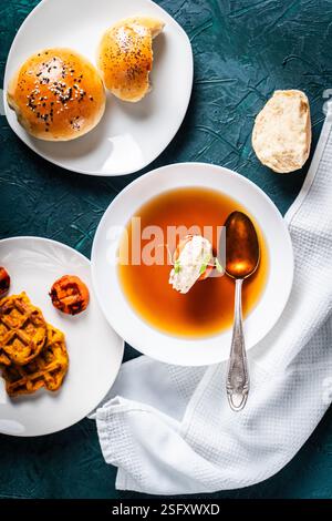 Suppe oder Consomme, dicke Fleischbrühe mit Karotten, Hähnchenknödel serviert auf einem weißen Teller auf dunkelgrünem Hintergrund mit Sesambrötchen, Serviette und Stockfoto